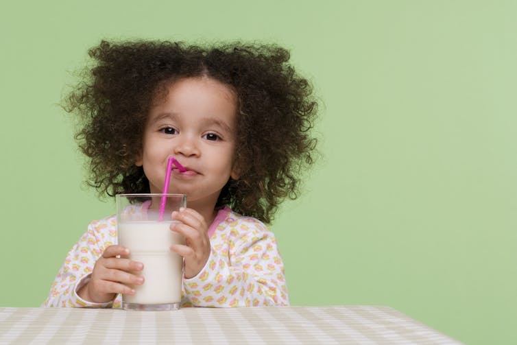 Una ragazza sorridente che beve un bicchiere di latte attraverso una cannuccia.