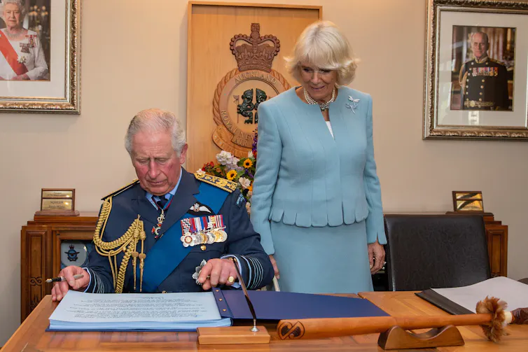 Prince Charles signs a guest book at a desk while Camilla stands to his left looking on