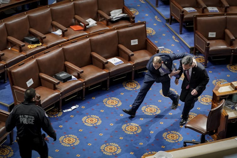 A police officer runs, ducking, with another man - both wearing black suits - across the U.S. House floor.