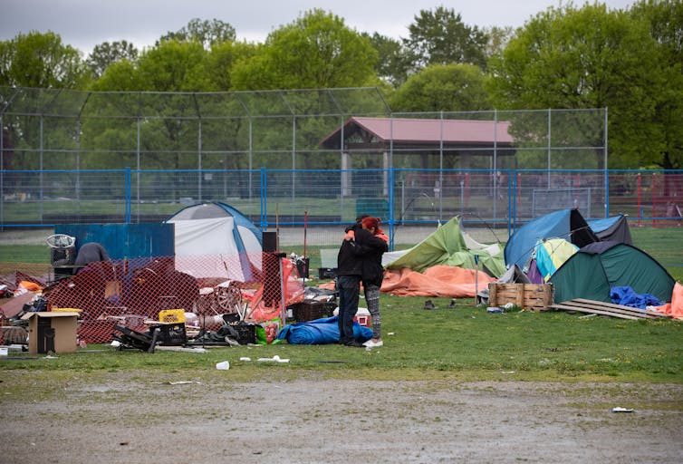 Two people hugging each other. In the background, tents, crates, sleeping bags, coolers, shoes and other personal belongings can be seen.