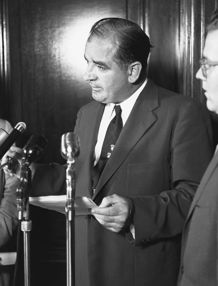 A man in a suit speaks into microphones in a black-and-white photo.