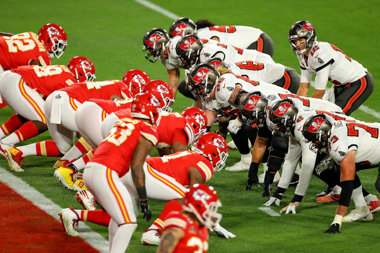 Linemen from two football teams, one in red uniforms and one in white, face off to await the snap.