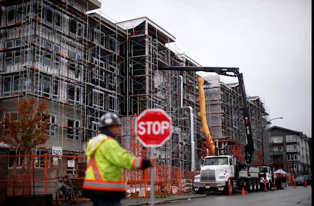 A construction worker in a bright yellow jacket and hard hat holds a stop sign in front of a condominium that's under construction