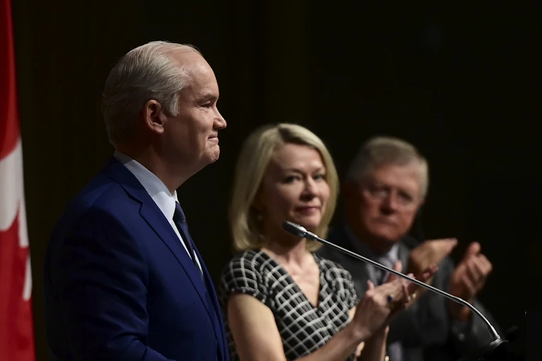 A man stands in front of a microphone in silhouette while a blonde woman looks into the camera beside him.
