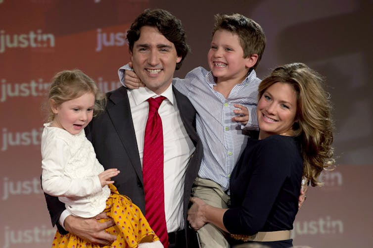 A man with dark hair, white shirt and red tie holds two children while a woman with dark blonde hair stands to his left.