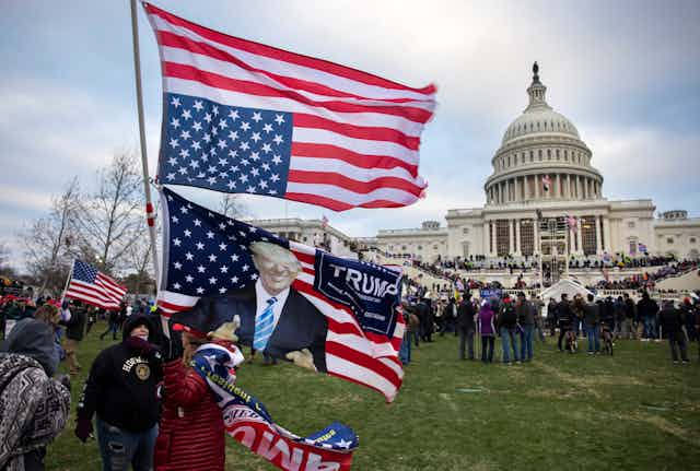 People hold up an upside down American flag and a flag showing Donald Trump, giving a thumbs-up, with crowds gathered in front of the U.S. Capitol.