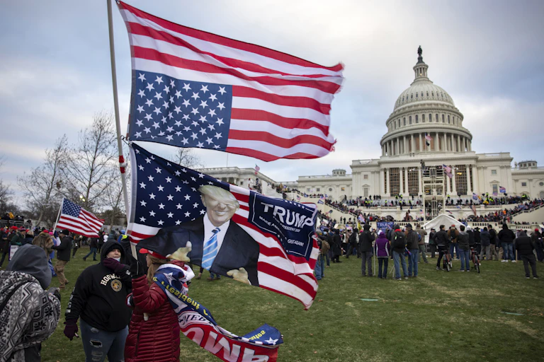 People hold up an upside down American flag and a flag showing Donald Trump, giving a thumbs-up, with crowds gathered in front of the U.S. Capitol.