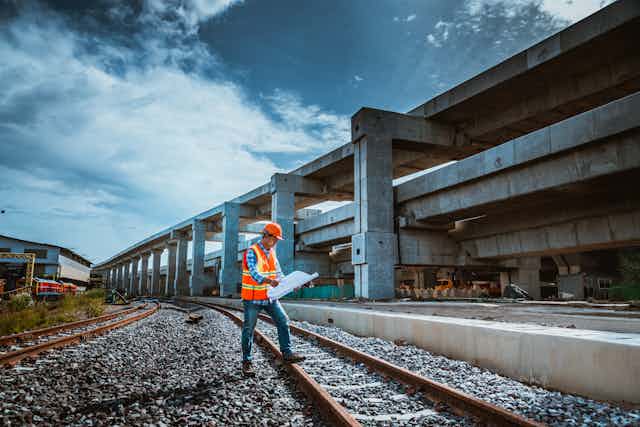 An engineer in orange vest and hard hat stands on a train track with an empty, grey building behind him.
