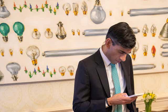 Rishi Sunak looking at his phone in front of a mural depicting lots of different lightbulbs. 