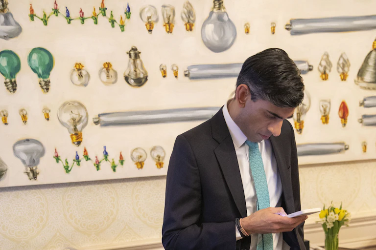 Rishi Sunak looking at his phone in front of a mural depicting lots of different lightbulbs.