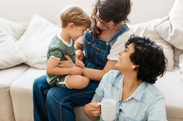A lesbian couple smiling at their young son, one of the women is seated on the couch with the child on her lap, the other is seated on the floor holding a cup of coffee