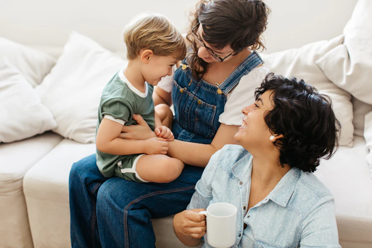 A lesbian couple smiling at their young son, one of the women is seated on the couch with the child on her lap, the other is seated on the floor holding a cup of coffee