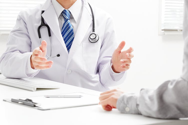 A doctor shown from the shoulders down, seated at a table and talking to a patient. The doctor has a stethoscope around his neck.