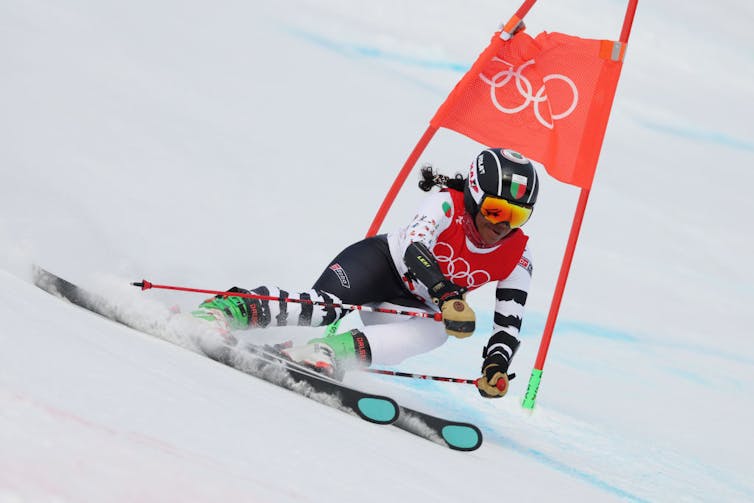 A woman with a ponytail in red and black outfit skis down a slope, cutting past a flag.