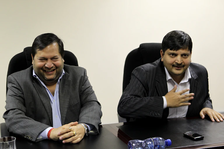 A wearing a jacket an open neck shirt laughs at a press conference while another, to his left, has his right hand on his chest to make a point.