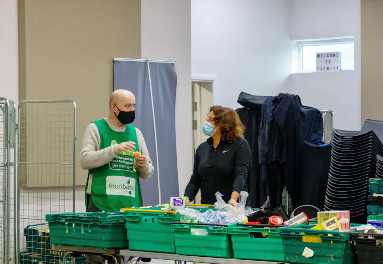 Man in apron reading 'food bank' talks to another volunteer as they organise food products at a table
