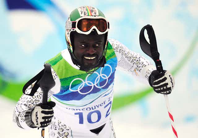 A male skier holds his skis and smiles, his goggles on his forehead and dressed in green, blue and white.