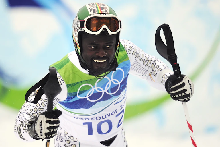 A male skier holds his skis and smiles, his goggles on his forehead and dressed in green, blue and white.