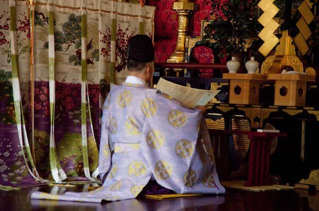 A Shinto priest wearing a religious white gown with gold motifs, sitting before an altar.
