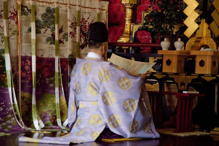 A Shinto priest wearing a religious white gown with gold motifs, sitting before an altar.