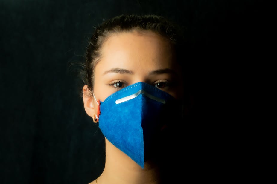 Person wearing blue face mask against black background looking directly into camera.