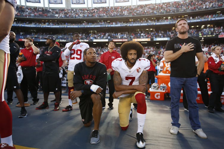 Two professional football players kneel on the field surrounded by teammates.