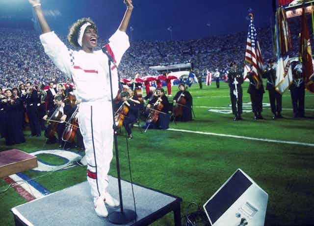 A black woman at a microphone extends her arms from a platform in the center of a football field, with an orchestra seated behind her.