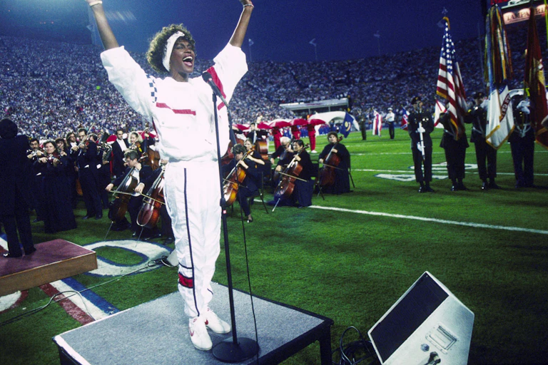 A black woman at a microphone extends her arms from a platform in the center of a football field, with an orchestra seated behind her.