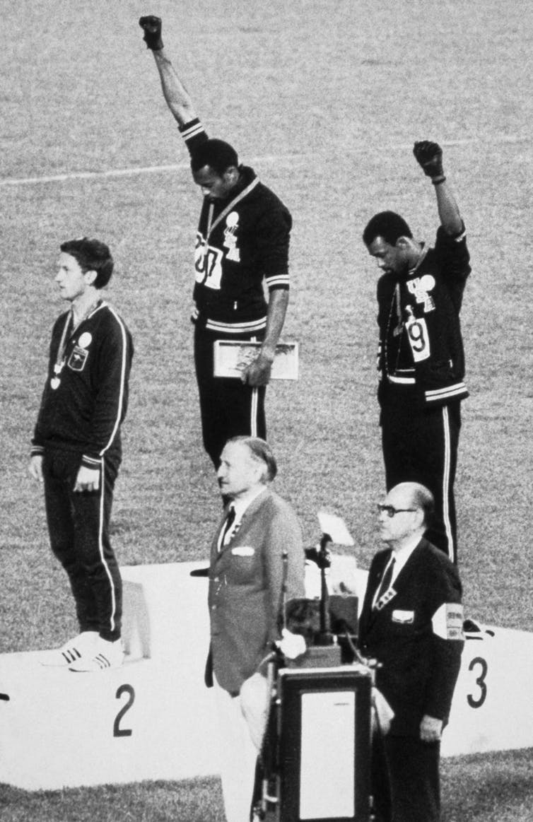 Tres atletas, uno blanco y dos negros, que llevan medallas se paran en un campo, con los hombres negros levantando sus puños con guantes negros.