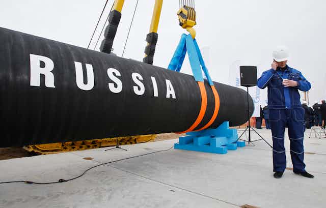 A piece of oil pipeline with the word RUSSIA written on it is held by a crane next to a worker wearing a construction hat alking on the phone