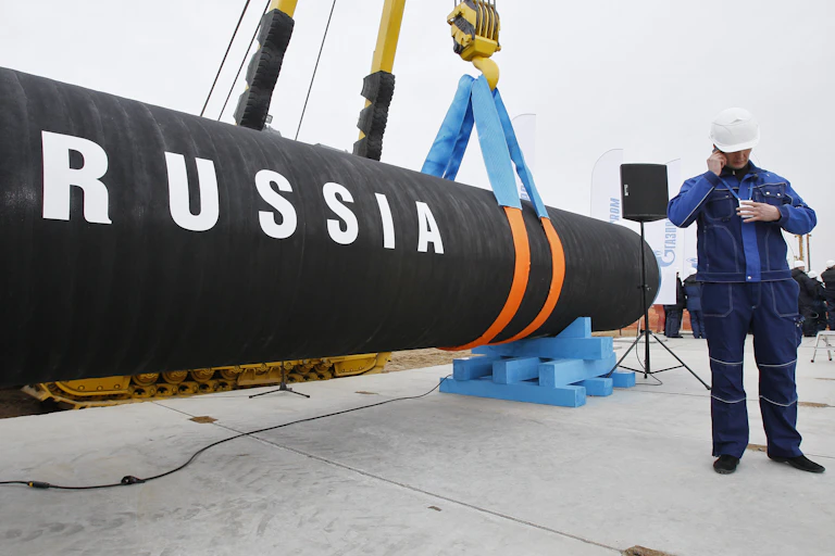 A piece of oil pipeline with the word RUSSIA written on it is held by a crane next to a worker wearing a construction hat alking on the phone