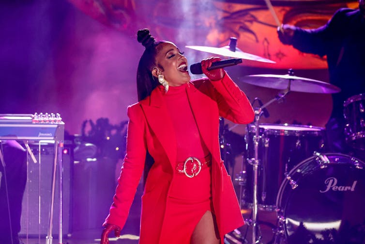 A Black woman dressed in a bright red jacket sings into a microphone.