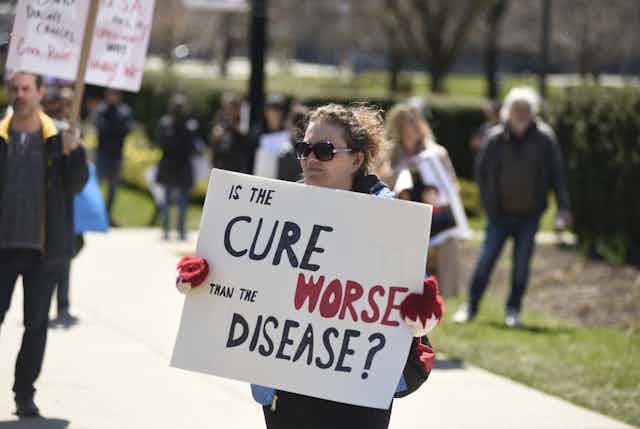 A woman holds a sign that reads 'is the cure worse than the disease?'