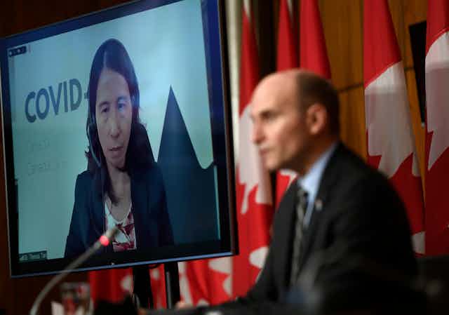A large video screen with an image of Chief Public Health Officer Dr. Theresa Tam, with Health Minister Jean-Yves Duclos in the foreground in front of a row of Canadian flags