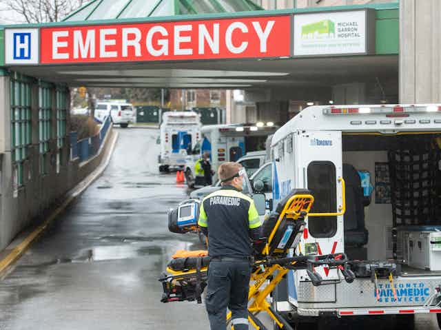 Paramedics in the foreground of a row of ambulances under a hospital's Emergency entrance