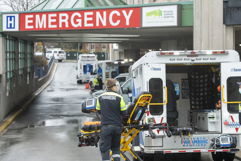 Paramedics in the foreground of a row of ambulances under a hospital's Emergency entrance