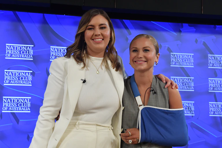 Brittany Higgins and Grace Tame arm in arm in front of press club backdrop.