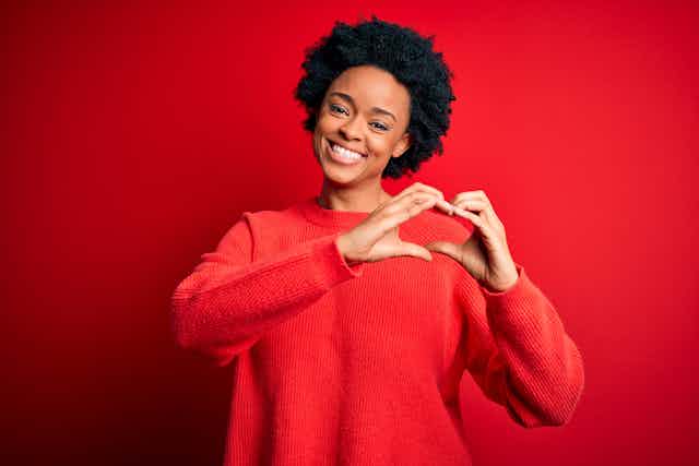 Young woman wearing red top, standing against red background, making a heart shape with her hands