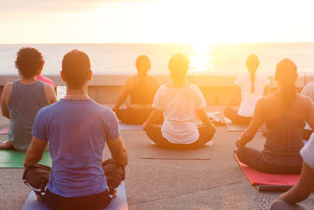 People sit crosslegged in a yoga pose, facing the ocean and the sun.