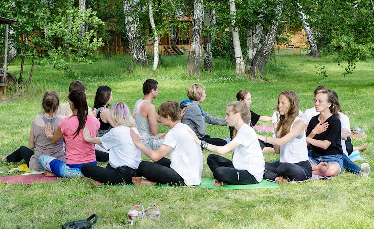 People sit in a circle holding each other's shoulders, in a green setting.