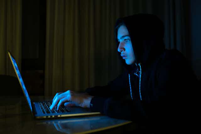 A teenage boy wearing a hoodie sits in front of a laptop in a dark room.