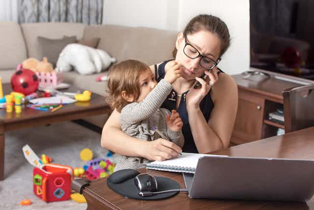 Young woman looking stressed talks on a mobile phone and takes notes in a notebook while holding her child on her lap. The room behind her is strewn with toys.