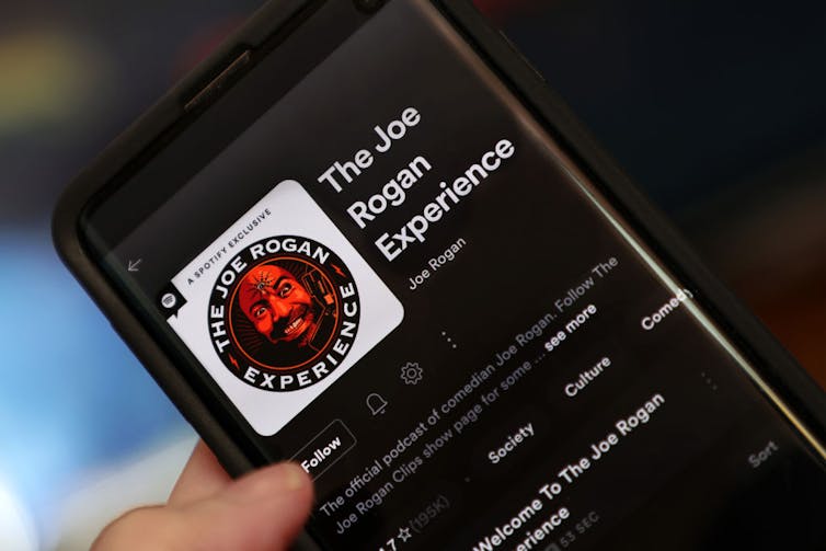 Closeup of hand holding smartphone with 'The Joe Rogan Experience' pulled up.