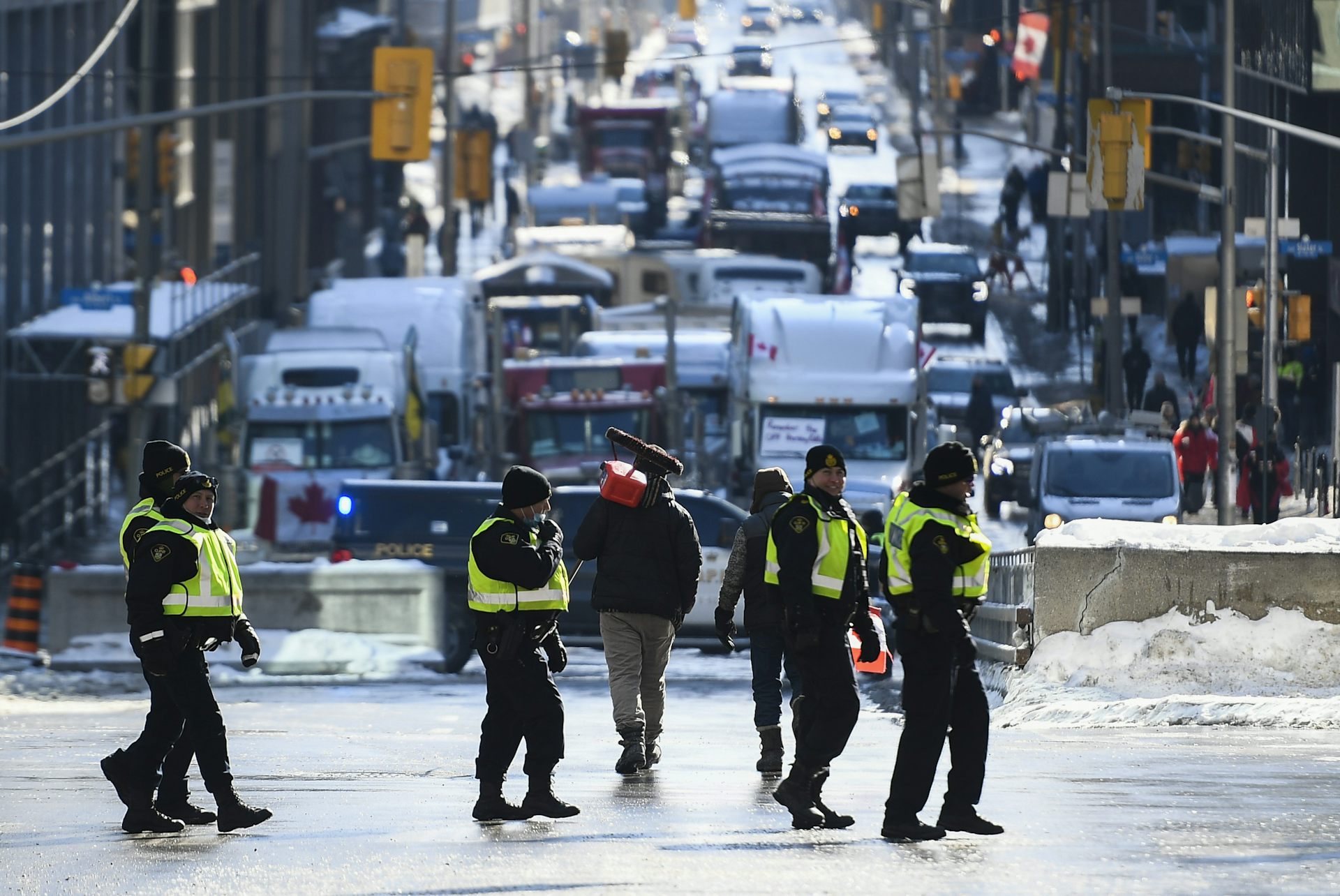 A protester carrying an empty fuel container on a broom handle walks past police officers toward parked transport trucks.