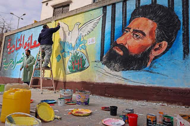 A person paints a mural showing a dove and a portrait of a bearded man in a cell