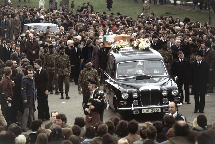 A hearse carries a coffin draped with the Irish flag through a crowd