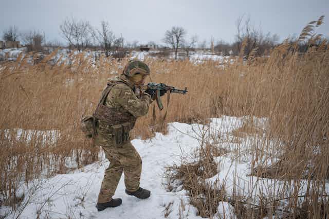 A soldier in battle fatigues and pointing his machine gun om front of him walks in a snowy field.