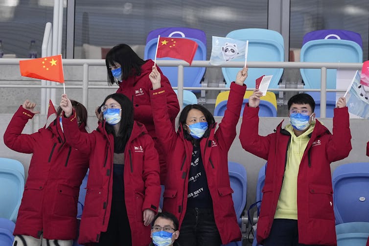 Four people in long, red winter coats wave Chinese flags at the Olympics