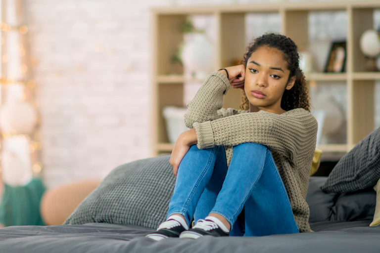 A teenage girl is sitting on her bed, looking unhappy.