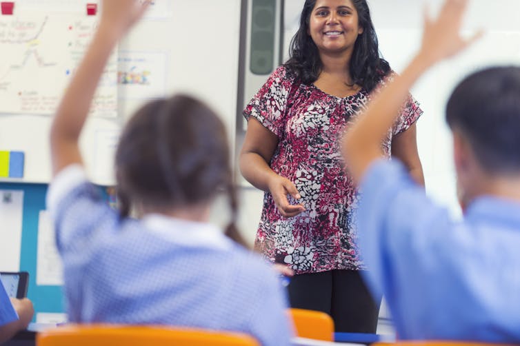 A teacher in a classroom with children you have their hands raised.